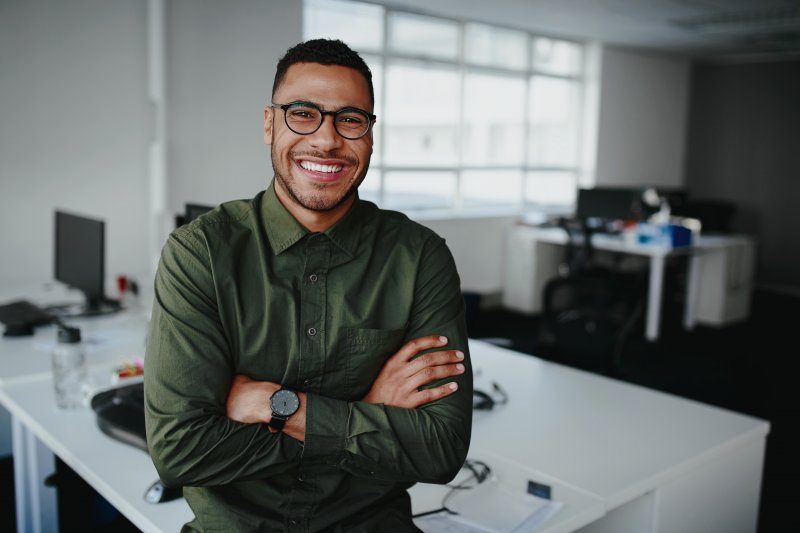 Man in dark green button up shirt smiling and standing with his arms crossed in an office